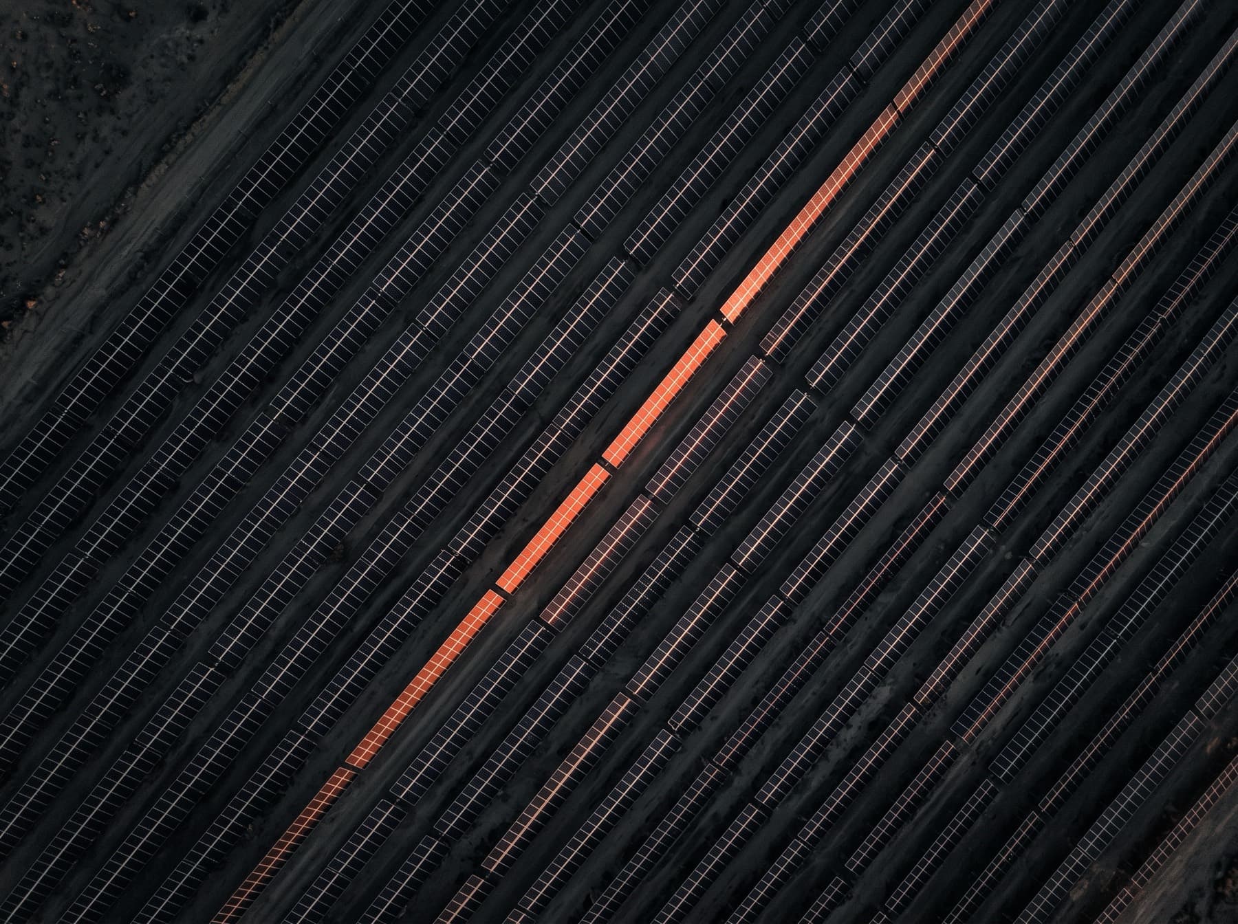 Aerial view of diagonal solar panel rows in near-darkness, one row catching an orange sun glint